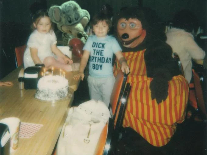 A vintage birthday party photograph showing early Chuck E. Cheese-era mascots, illustrating the character's long history before its landfill-photo revival.