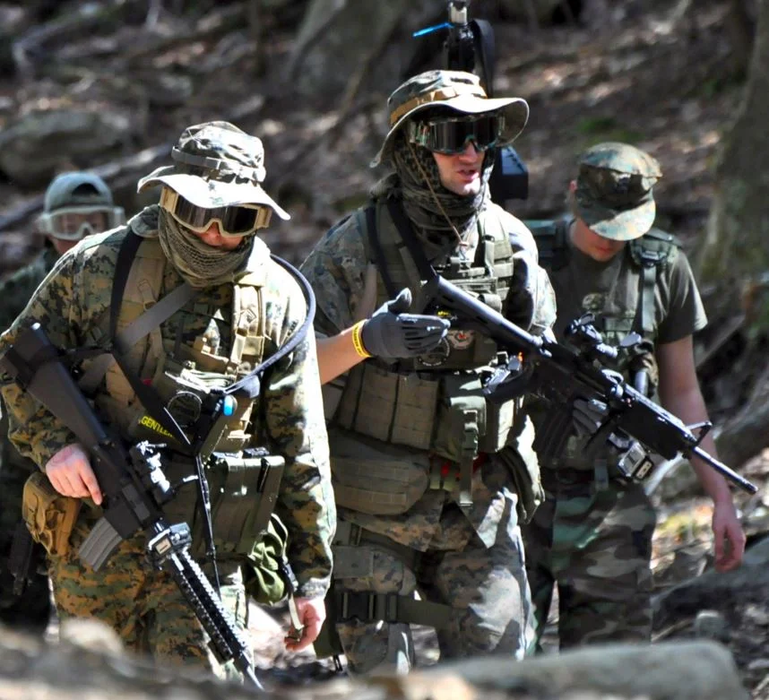 Airsoft players geared up in tactical camouflage and protective equipment during a woodland skirmish.