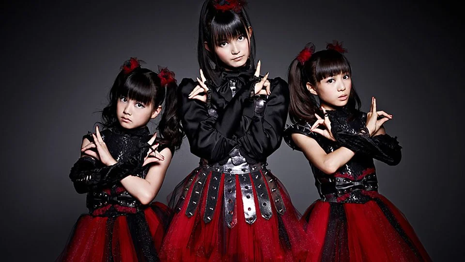 The three members of Babymetal in their iconic black-and-red stage costumes striking their signature poses in an official promotional photo.