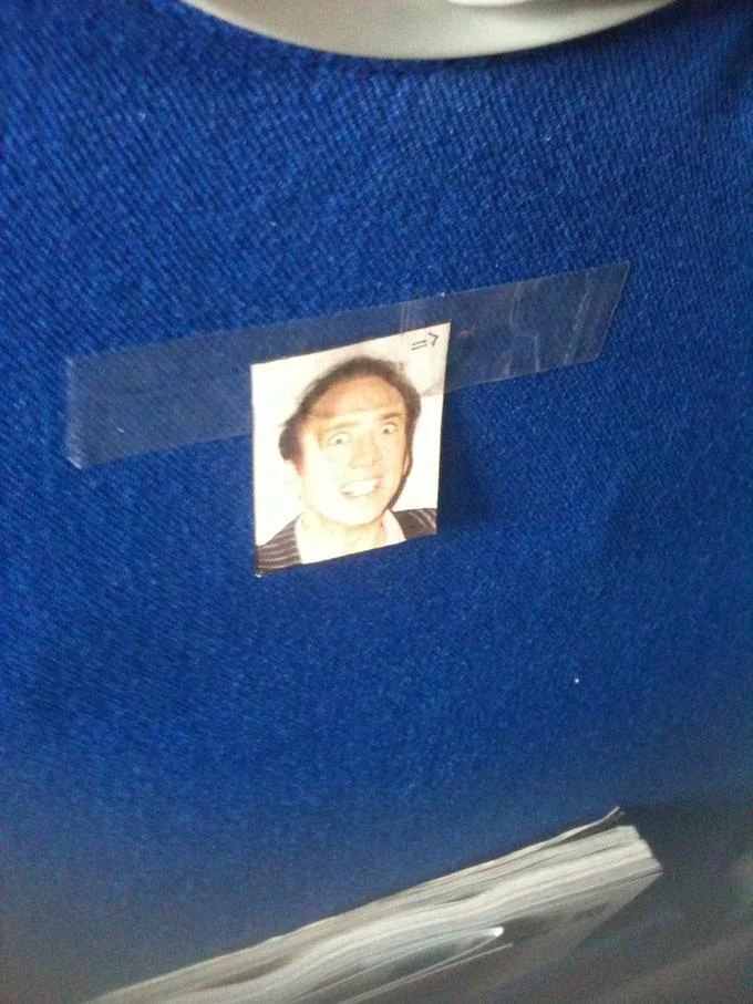 A miniature Nicolas Cage portrait taped to a public transit seat as a low-key caging in the wild.