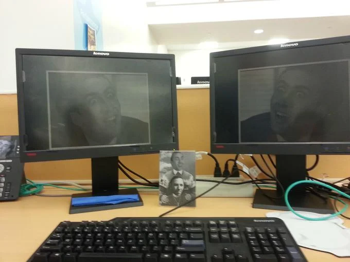 A workplace caging: an office desk with dual monitors set to Nicolas Cage screensavers and a framed portrait of the actor.