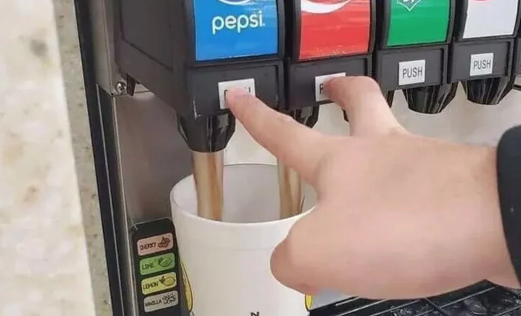 The original photo behind the 'Double Soda Pour' meme, showing a hand simultaneously dispensing two rival sodas into the same cup.