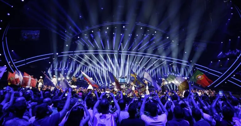 The Eurovision Song Contest live arena, with fans waving flags of participating countries in front of the illuminated main stage.