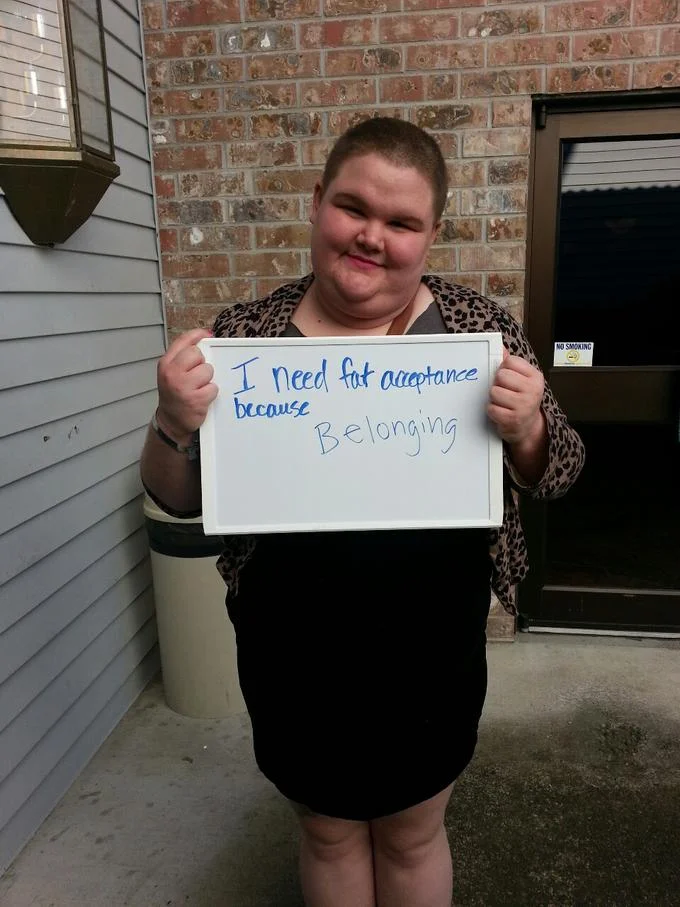 A participant in the 'I Need Fat Acceptance Because' photo campaign holds a sign citing 'Belonging' as their reason for supporting the movement.