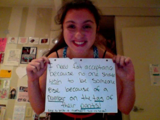 A participant in the 'I Need Fat Acceptance Because' campaign holds a handwritten sign explaining why body size shouldn't dictate self-worth.