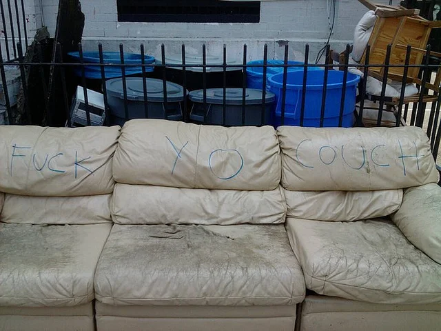 A trashed sofa in an alley beside a graffiti wall, spray-painted with the meme phrase 'Fuck Your Couch'.