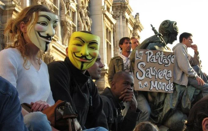 Anonymous-affiliated demonstrators wearing Guy Fawkes masks at a protest in Paris beside a sign reading 'Sharing will save the world.'