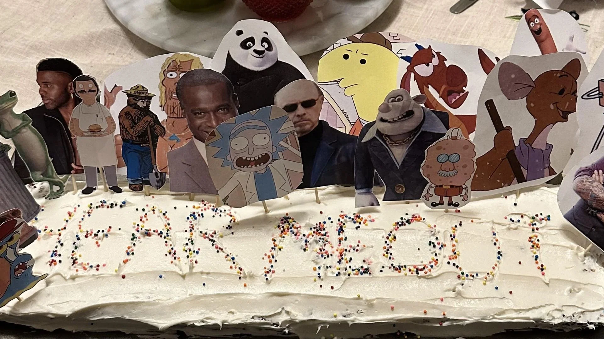 A 'Hear Me Out' cake featuring paper cutout toppers of unconventional fictional character and celebrity crushes, exemplifying the viral trend where participants defend their unusual attractions.