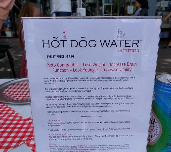 The Hot Dog Water product sign listing exaggerated health claims and a $37.99 event price, designed to parody wellness marketing.