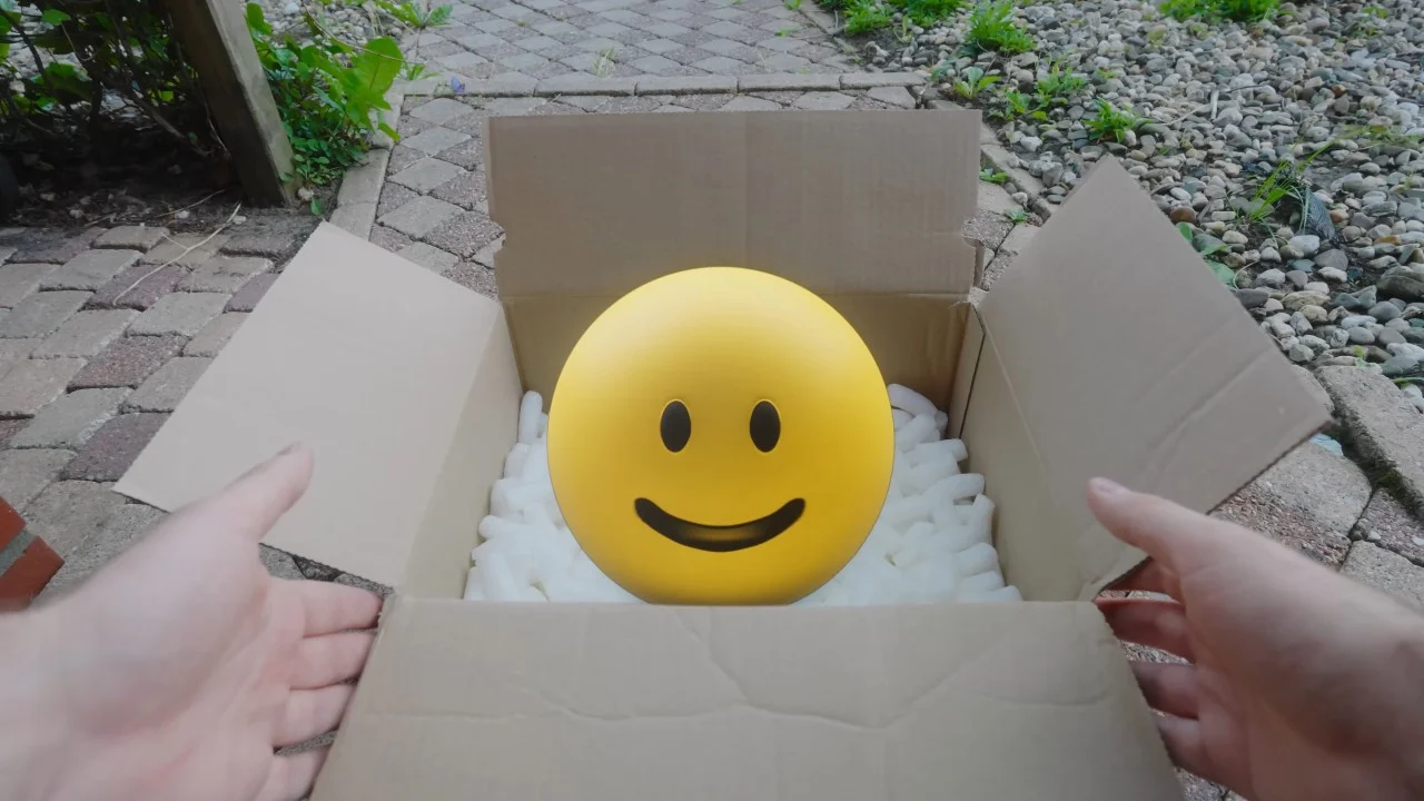 The signature unboxing shot: a smiling yellow emoji face 'Verity' arrives in a cardboard package.