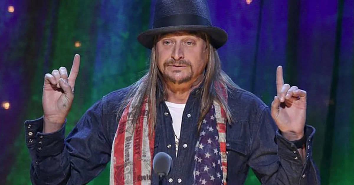 Kid Rock performing on stage in his signature fedora and American flag scarf.