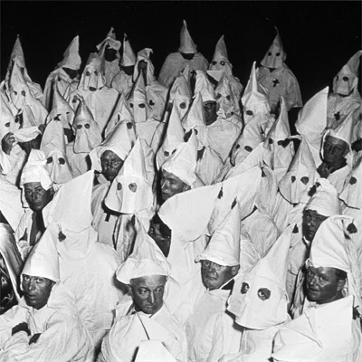 A historical black-and-white photograph of a Ku Klux Klan gathering, showing members in their iconic white robes and conical hoods.