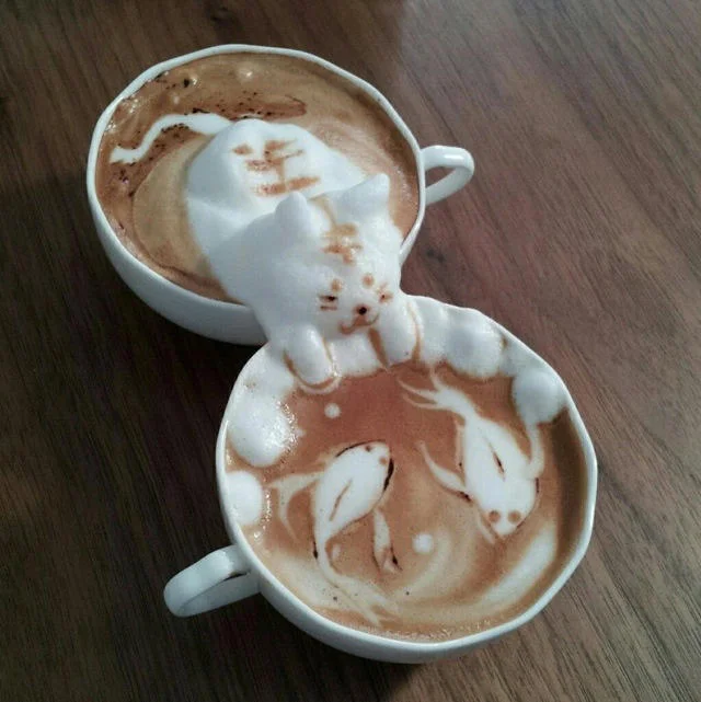 An elaborate 3D latte art piece featuring a foam-sculpted cat resting between two cups, with koi fish swirling in the coffee below.