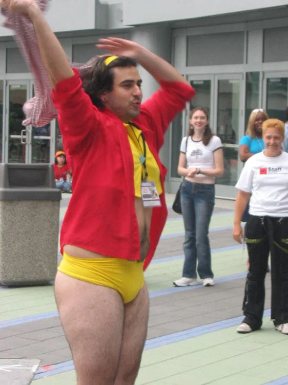 A Man-Faye cosplayer poses confidently in the classic yellow-and-red outfit at an anime convention.