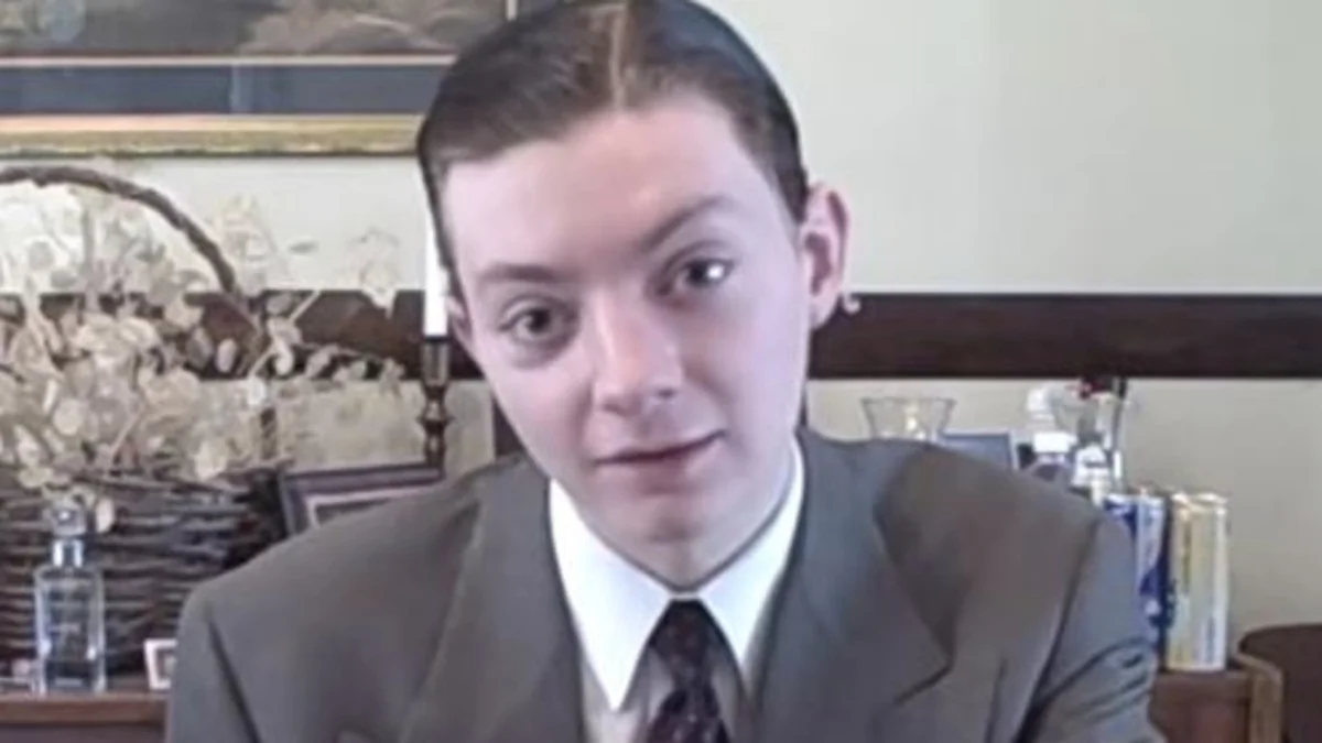 TheReportOfTheWeek's signature on-camera look: a young man in a formal suit with slicked-back hair, filming food and energy drink reviews from his home.